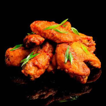 Fried chicken wings in spicy red glaze with sliced green onion, stacked on a black reflective surface