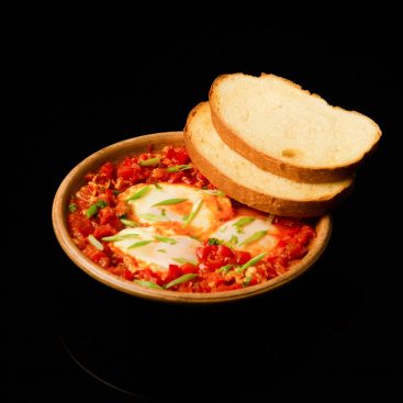 Shakshuka with eggs in tomato and pepper sauce, topped with green onion, served with white bread slices