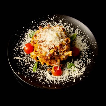 Tagliatelle bolognese with cherry tomatoes, basil and grated parmesan on a black plate, shot from above