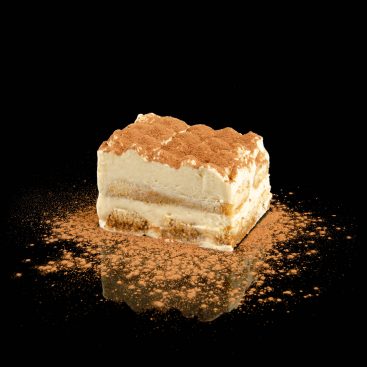 Tiramisu slice with cream layers, ladyfingers and cocoa dusting on a black reflective surface