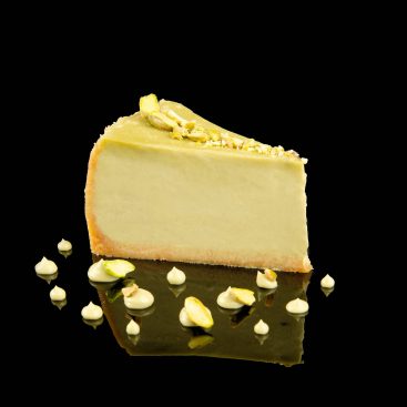 Slice of pistachio cheesecake topped with chopped pistachios, with dots of cream, shot from the side